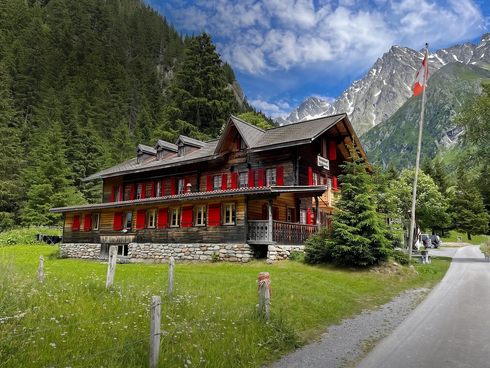 Das Haus von der Naturstrasse: drei Stockwerke Holz, rote Läden, Fahne, dahinter der Berg