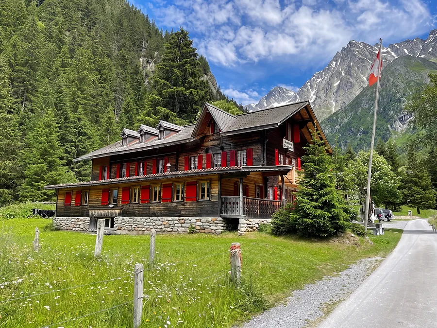 Das Haus von der Naturstrasse: drei Stockwerke Holz, rote Läden, Fahne, dahinter der Berg